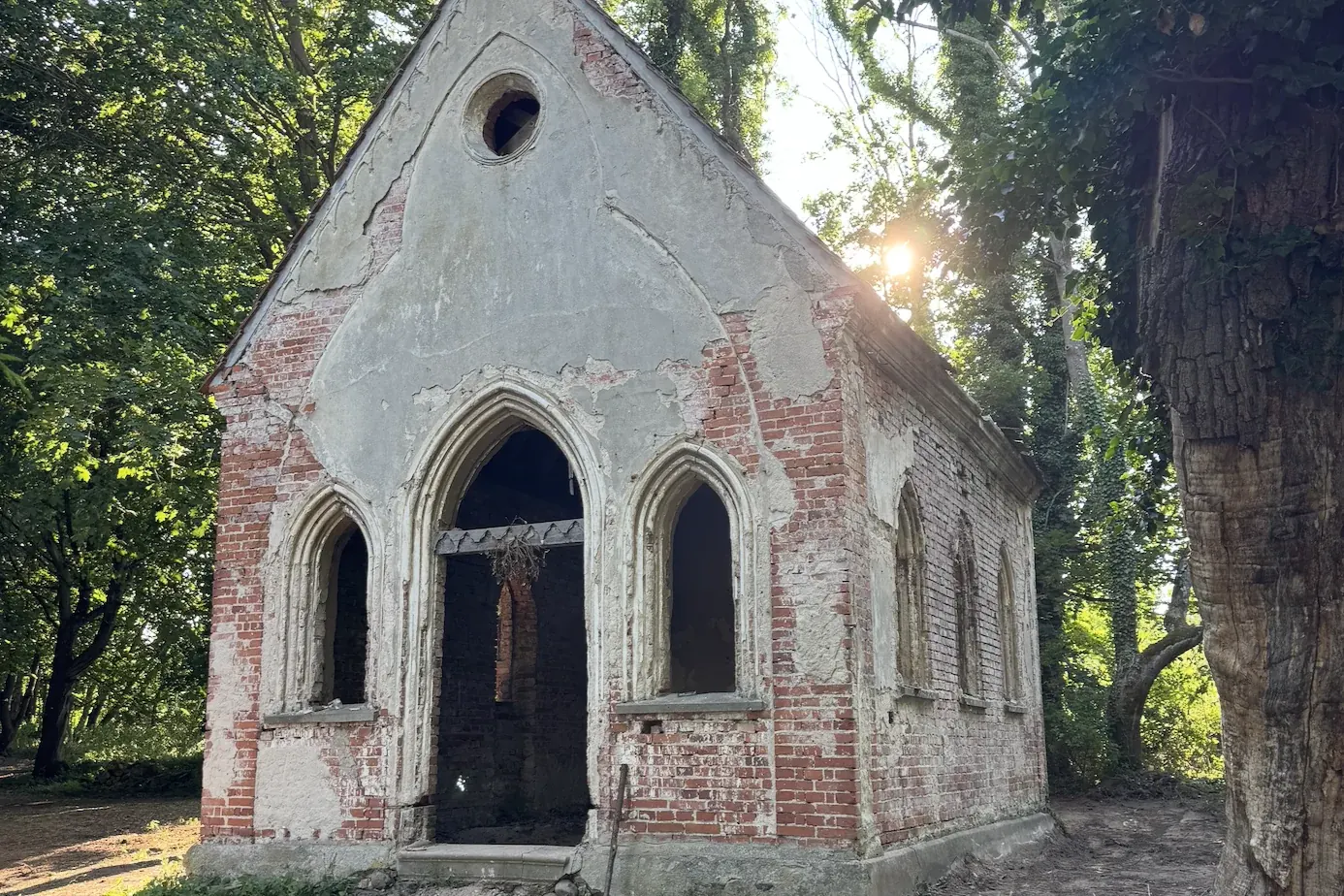 Die neugotische Kapelle von Rothspalk im Park: Backstein-Giebel mit Spitzbogenfenstern und Rundfenster, Dach und Putz beschädigt, Abendsonne durch die Bäume
